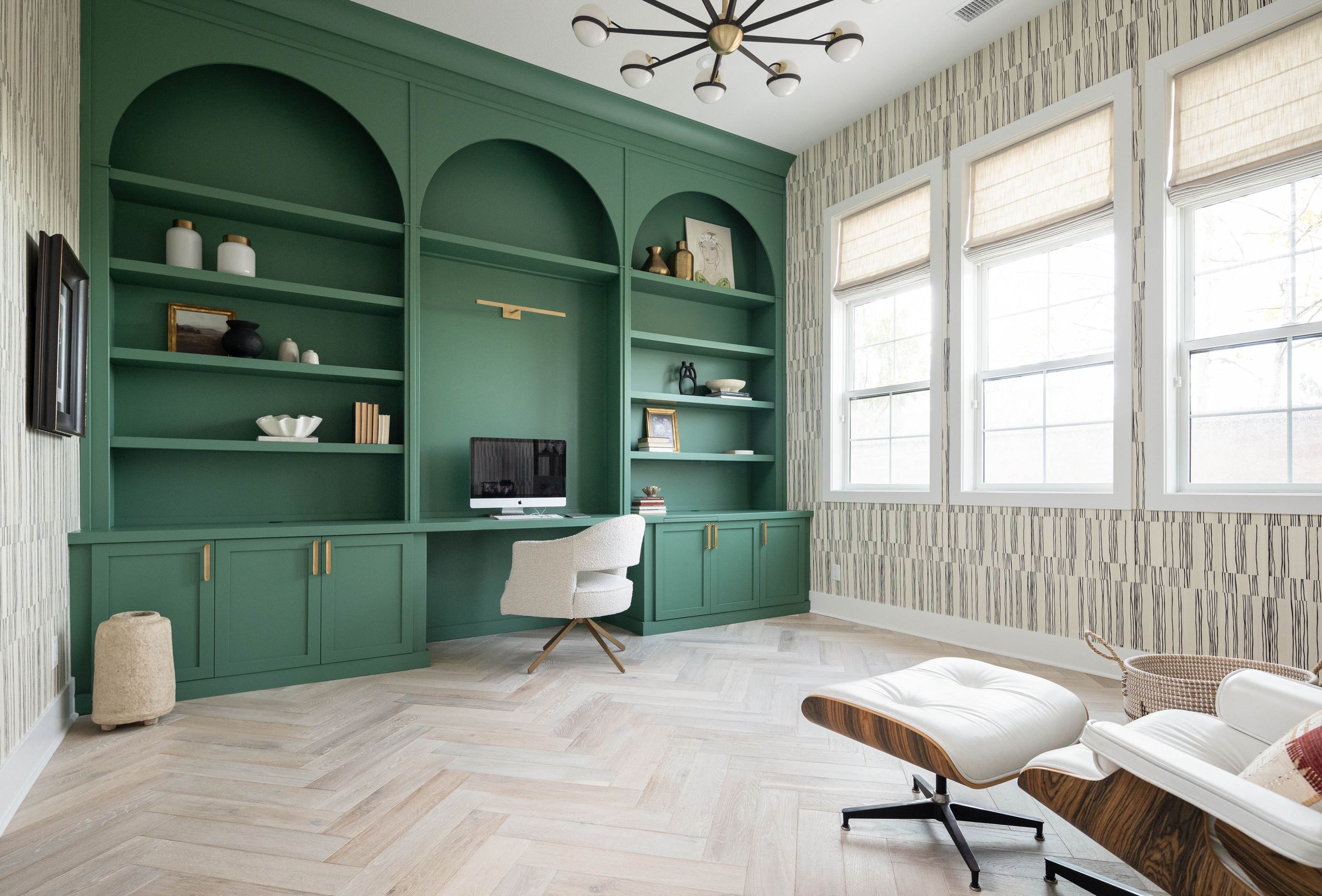 A home study with deep green arched built-in shelving, brass hardware, grasscloth wallpaper and pale herringbone floors.
