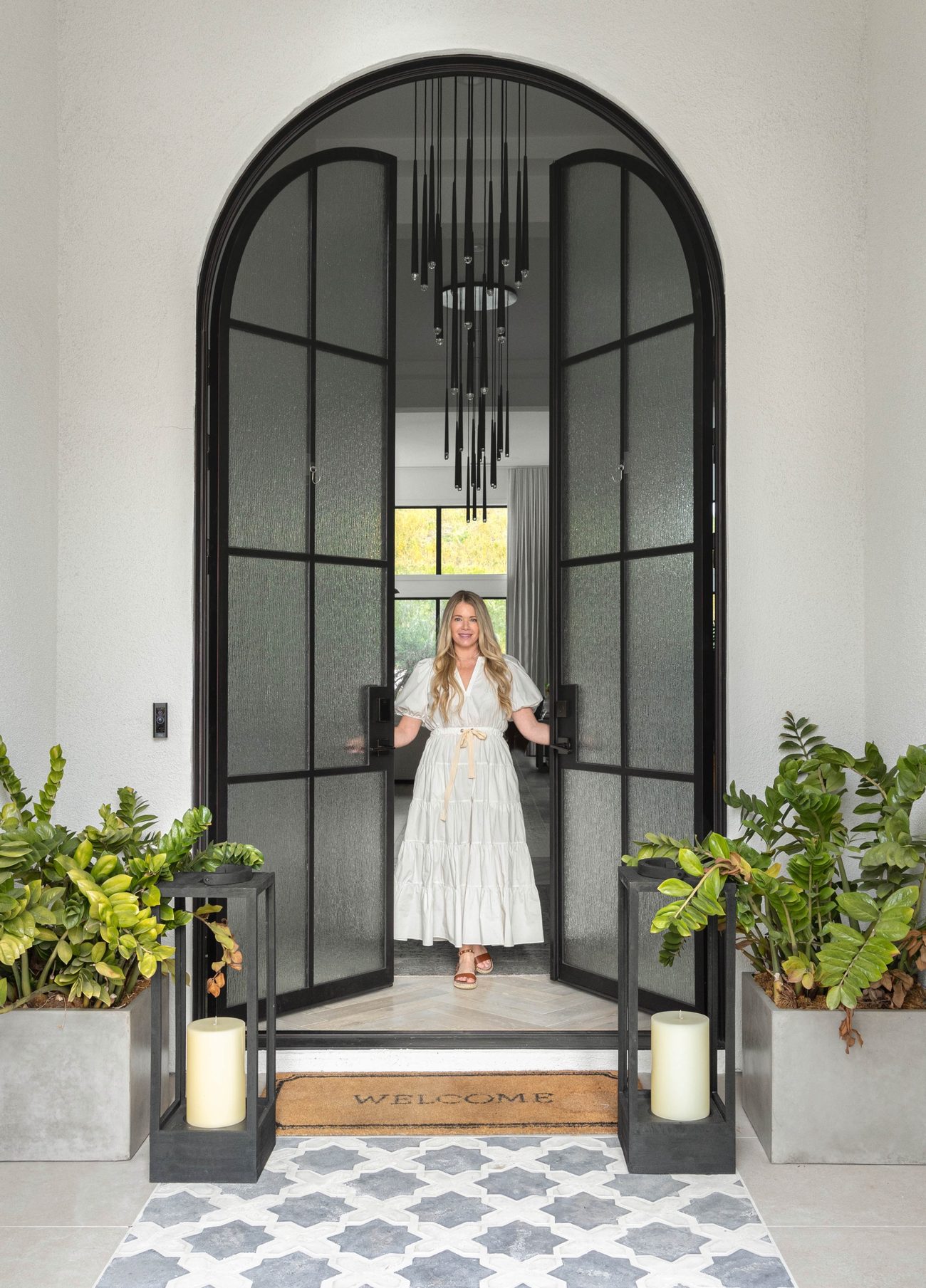 Kimberley Lustig opening a tall black steel arched entry door, with potted greenery and lanterns on the step.