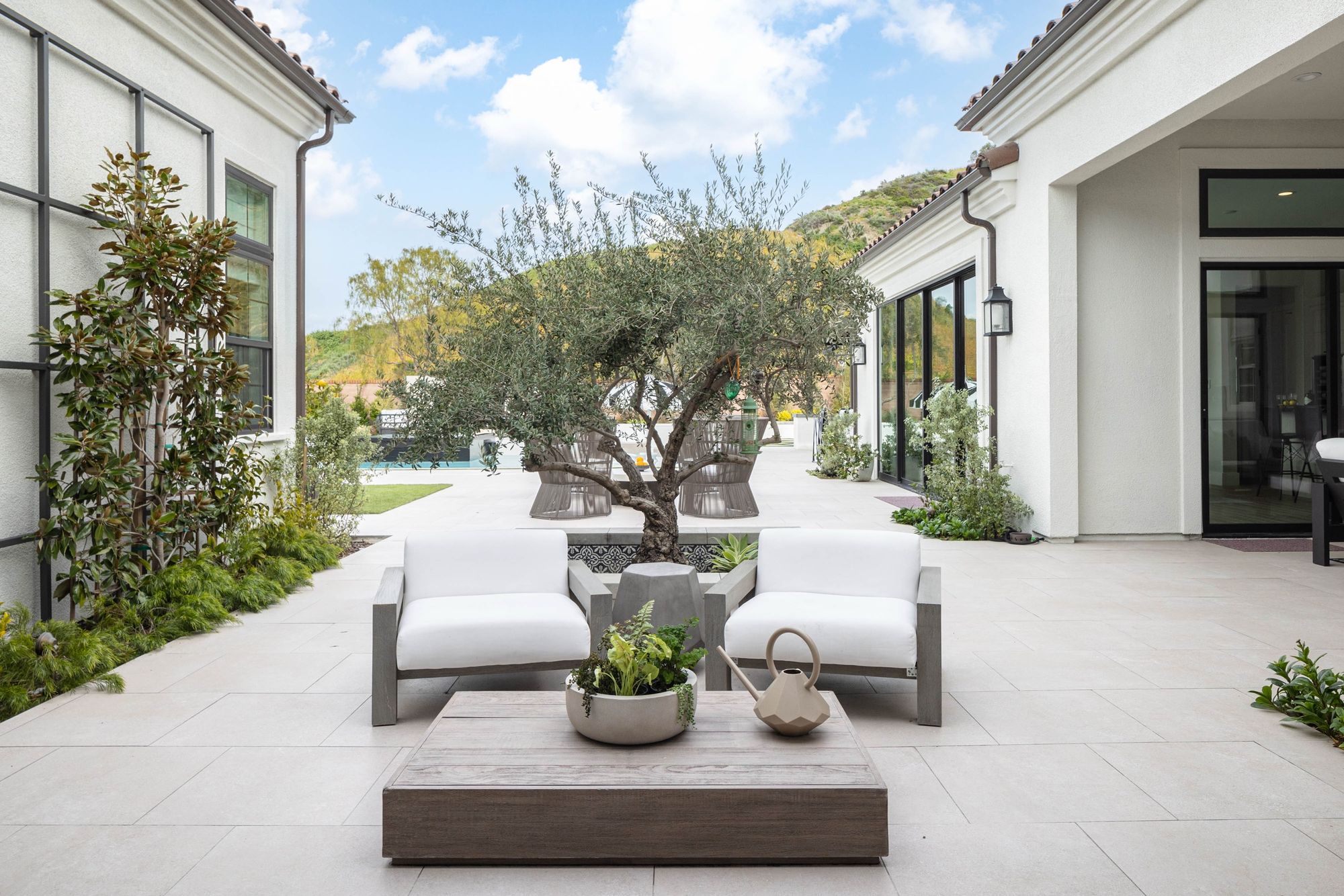 A limestone courtyard with teak lounge chairs set around a mature olive tree, a pool and hillside behind.