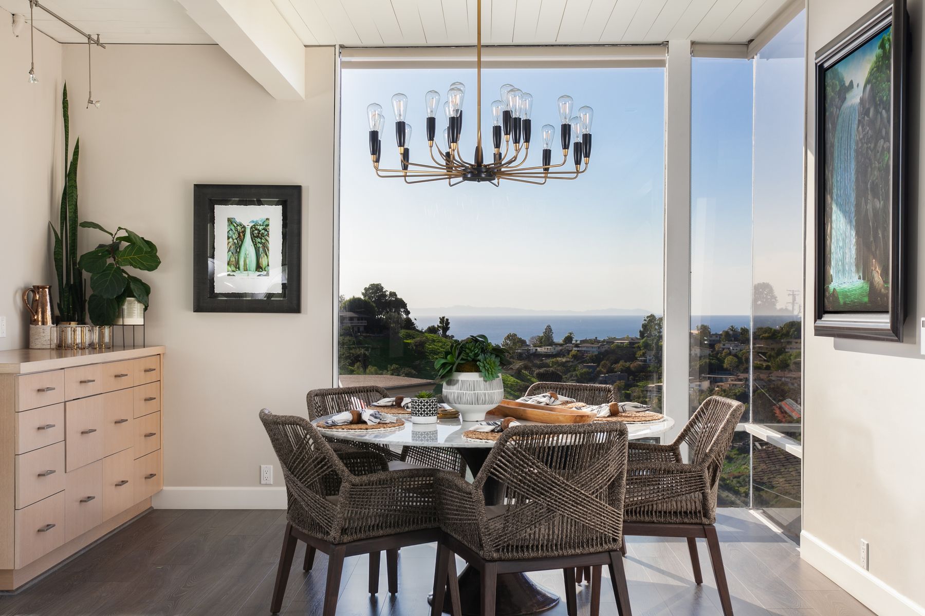 A dining nook with woven rope armchairs around a marble table, framed by a corner window over the coastline.