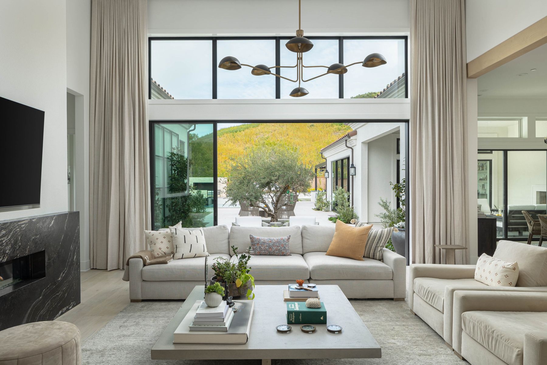 A double-height great room with ivory sectionals and full-height drapery, opening through a wall of glass onto a courtyard and green hillside.