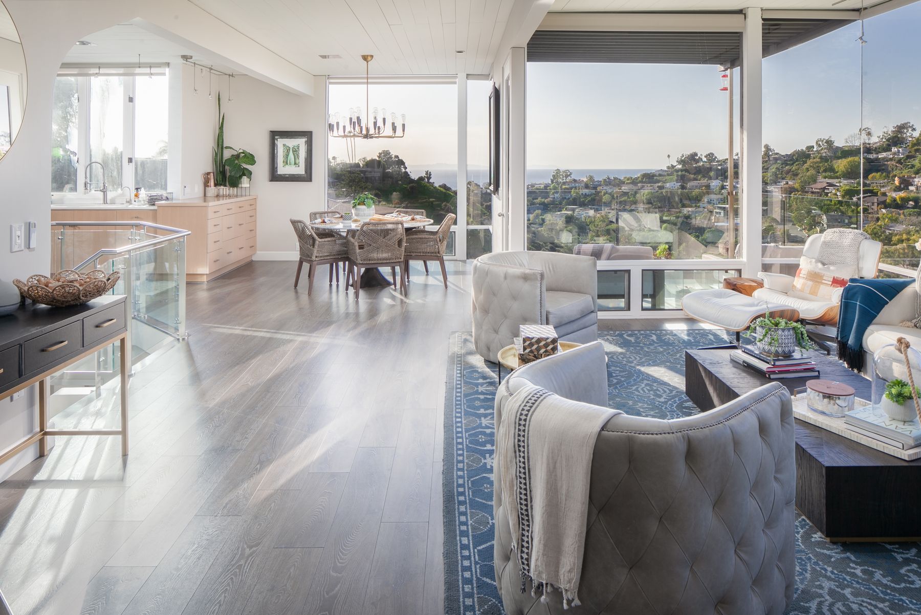 An open living and dining space with tufted swivel chairs, a blue patterned rug and walls of glass opening to an ocean view.